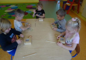 Pięcioro dzieci siedzi przy stole, w rękach trzymają gumki recepturki, które zakładają na kartonowe małe pudełko.